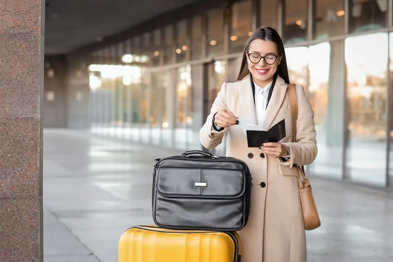 Frau mit Brille hält im Bahnhof ihr Ticket und ihren Ausweis, neben ihr Koffer und Reisetasche